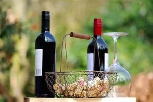 Two red wine bottles beside a wire basket of used corks and an upturned wine glass outdoors