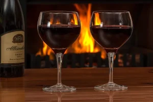 Two glasses of red wine and a bottle on a wooden table in front of a fire