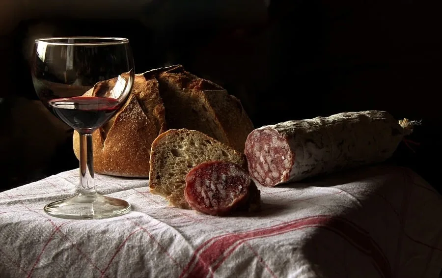 Glass of red wine beside a rustic bread loaf and cured sausage on a linen cloth