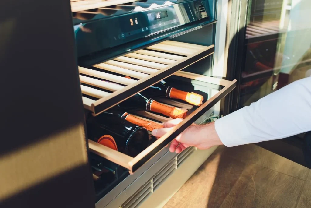 A hand pulls out a wooden shelf of bottles stored on their sides in a wine refrigerator