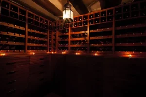 Dim wine cellar lined with wooden bottle racks, drawer cabinets and candles under a hanging lantern