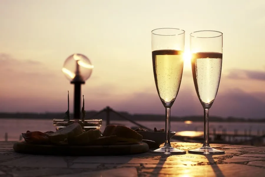Two flutes of sparkling wine on a waterside table at sunset with a snack platter