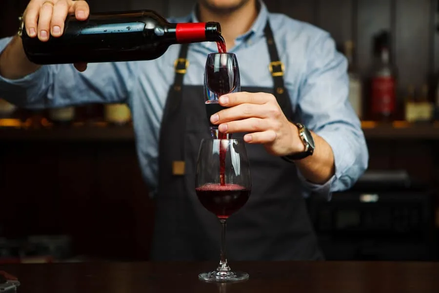 A sommelier pours red wine from a bottle through a handheld aerator into a glass on a bar
