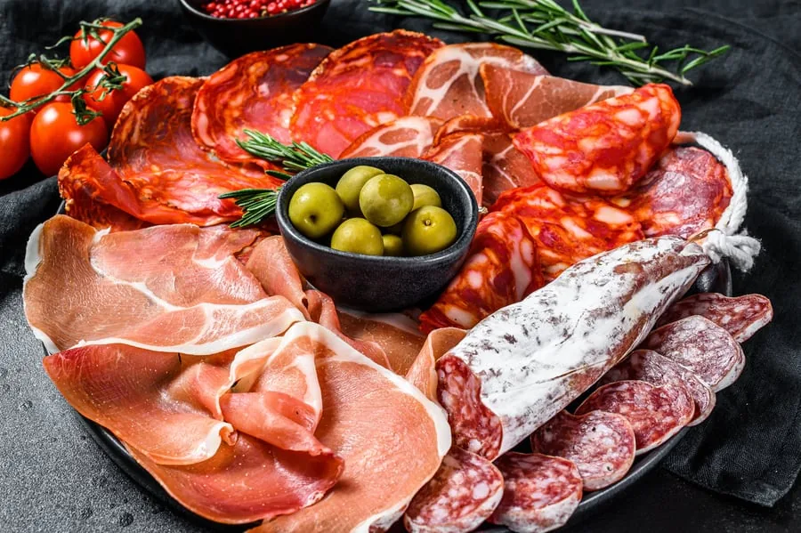 Platter of cured meats with sliced salami, dry sausage and thin ham beside a bowl of green olives