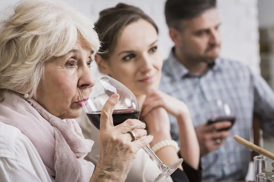 An older woman sips red wine from a glass while two others watch at a tasting