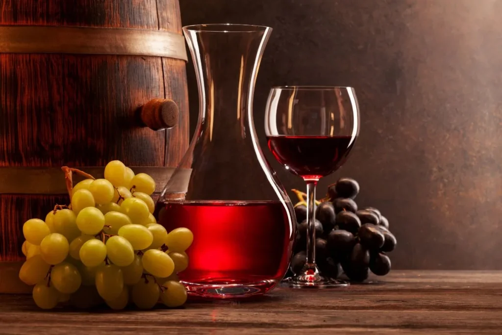 Decanter of red wine and a filled wine glass beside green and black grapes near an oak barrel