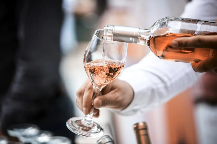 A server in a white shirt pours rose wine into a stemmed glass held by a guest