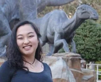 A smiling woman with long dark hair standing in front of a dinosaur statue outdoors