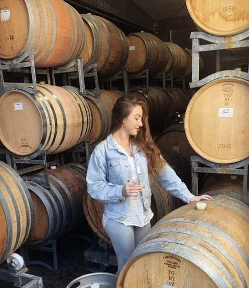 A woman holding a glass of wine among stacked oak barrels in a winery barrel room