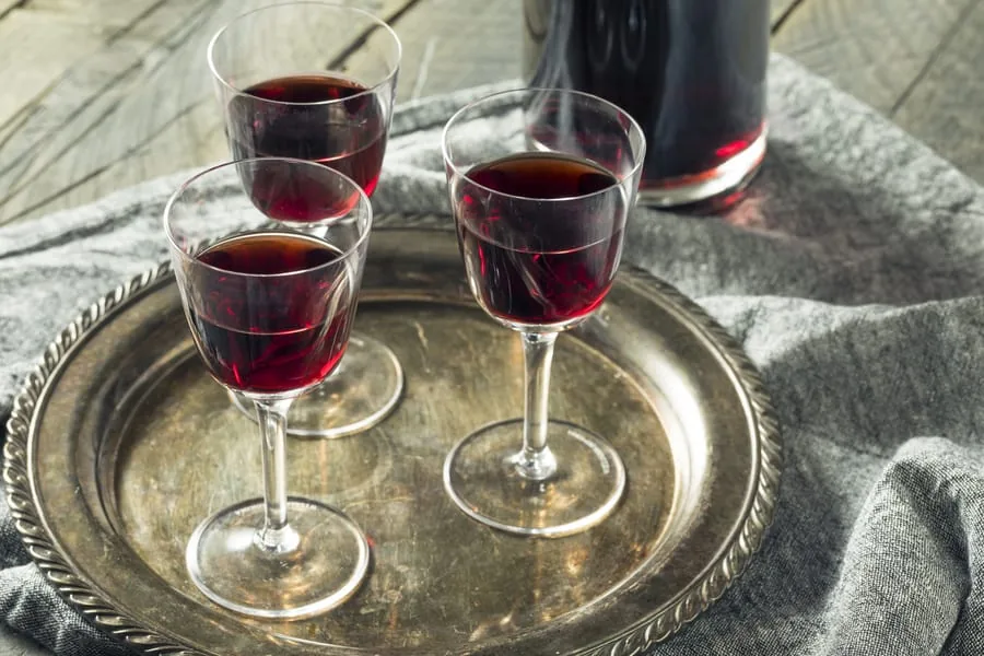 Three small stemmed glasses of dark red fortified wine on a metal tray beside a filled decanter