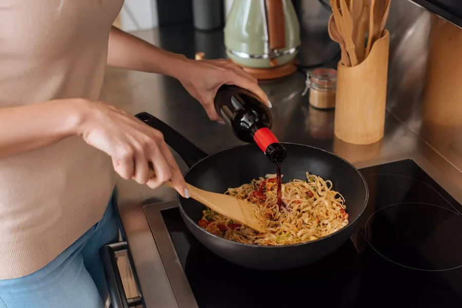 A woman pours red wine from a bottle into noodles cooking in a pan on the stove
