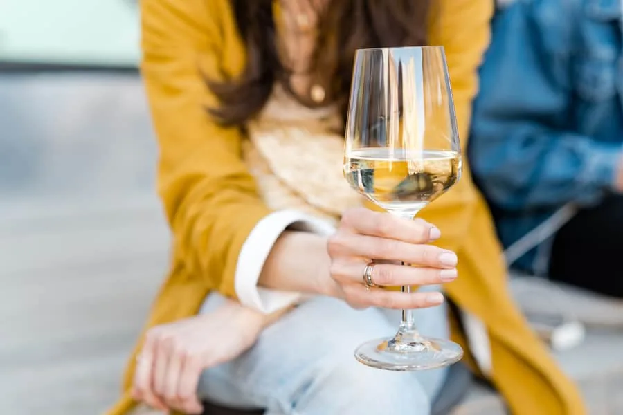 A woman in a mustard jacket holds a glass of chilled white wine while seated outdoors
