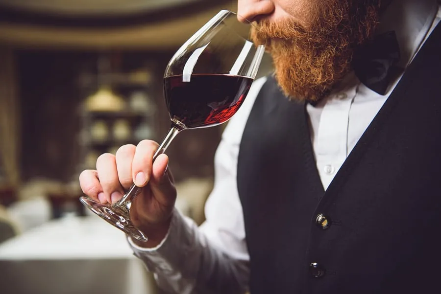 A bearded sommelier in a vest and bow tie smells red wine in a large tasting glass