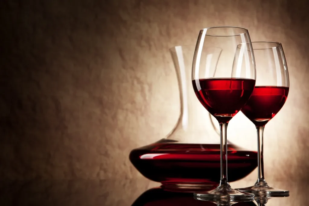 Two long-stemmed glasses of red wine beside a filled decanter on a reflective surface