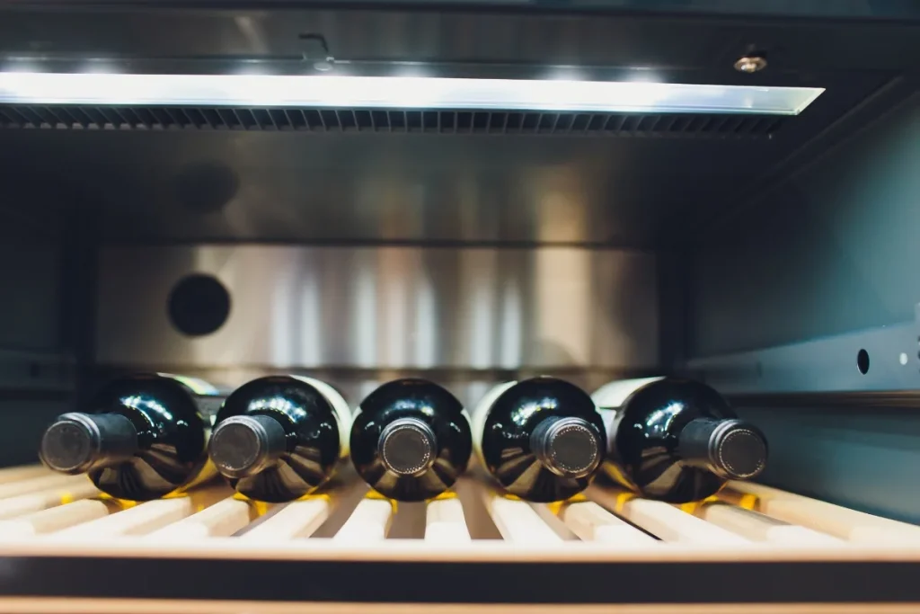 Five dark wine bottles resting on a wooden shelf inside a lit wine cooler