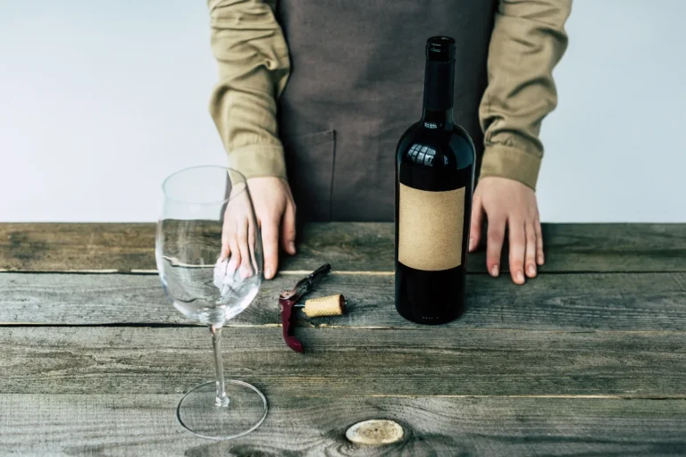 Opened wine bottle with a blank label beside a corkscrew, loose cork and empty glass on a wooden table