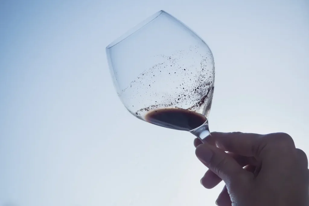 A hand tilts a nearly empty wine glass, showing dark sediment specks clinging to the bowl