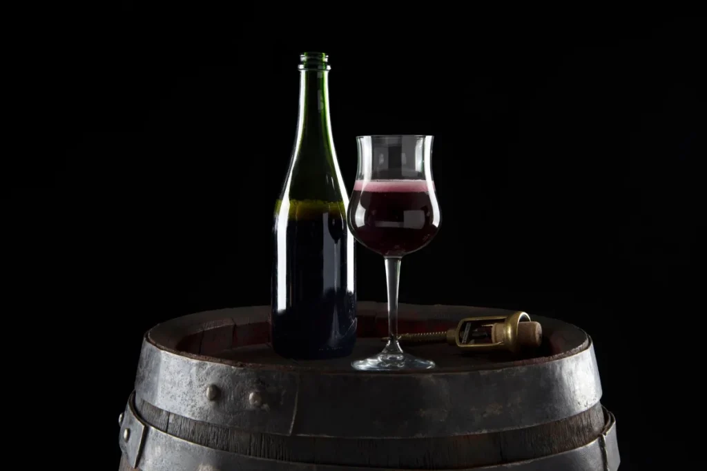 Opened bottle of red wine beside a filled glass and corkscrew on an oak barrel