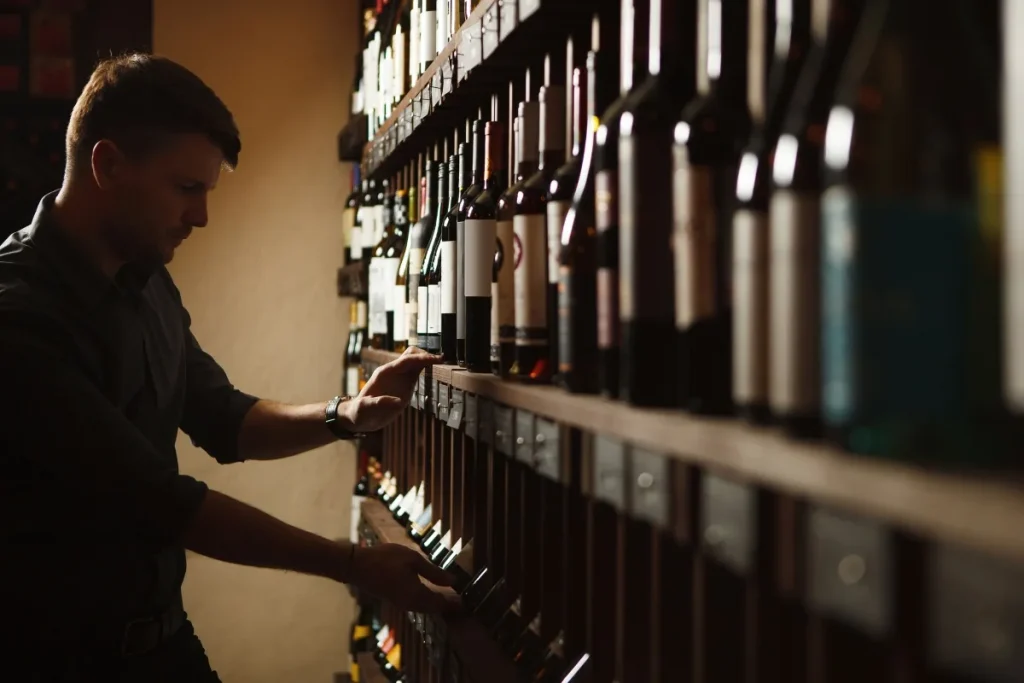 A man reaching for a wine bottle on tall wooden shelves in a dimly lit cellar