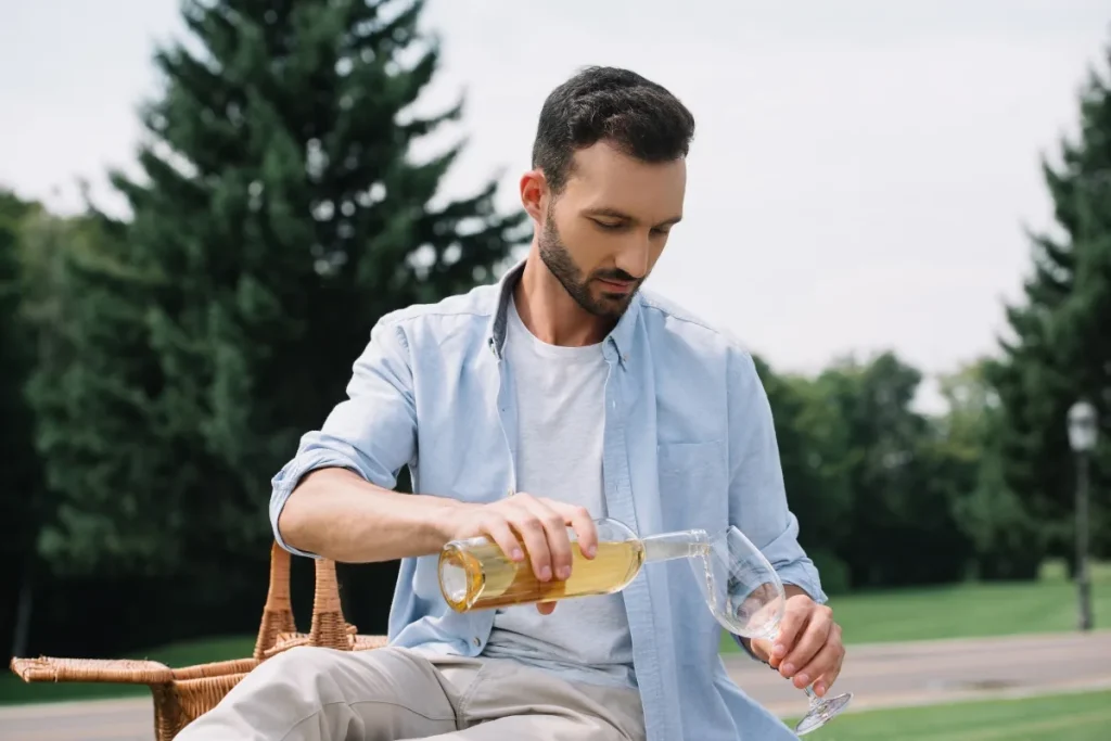 A man pours white wine from a clear glass bottle into a wine glass at an outdoor picnic