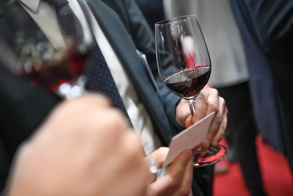 A hand holds a glass of red wine with tasting notes card at a crowded wine tasting