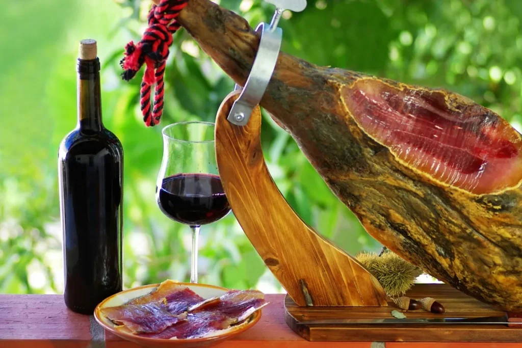 Bottle and glass of red wine beside a cured ham leg on a wooden holder with sliced ham on a plate