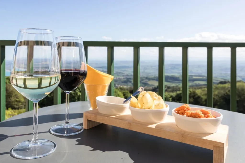Glasses of white and red wine beside bowls of chips and snacks on a sunny hillside terrace