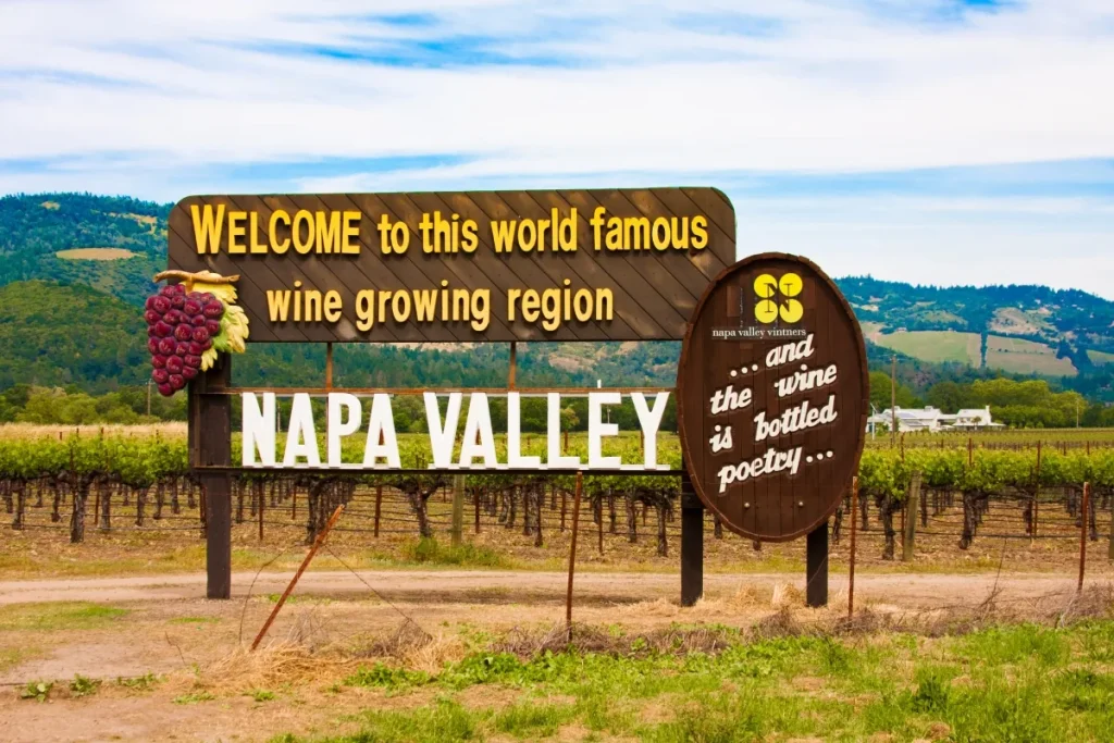 Wooden Napa Valley welcome sign beside vineyard rows with green hills behind