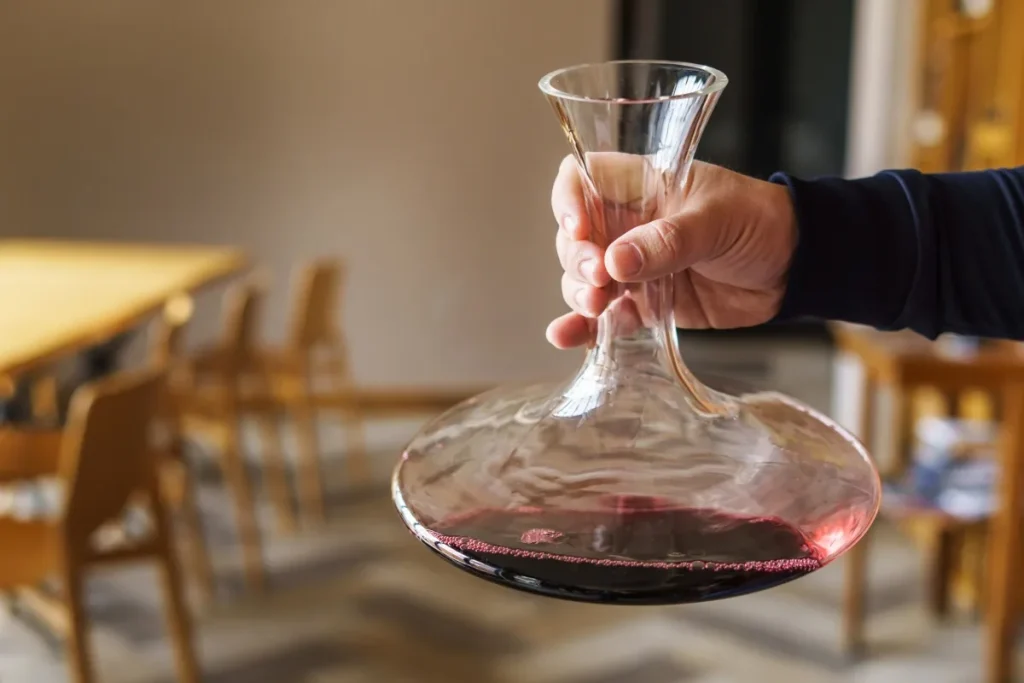 A hand swirling red wine in a wide glass decanter above a dining room floor
