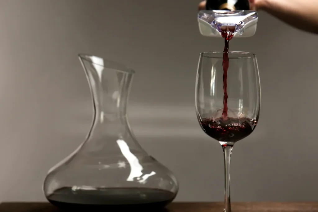 Red wine poured through a handheld aerator into a glass beside a glass decanter
