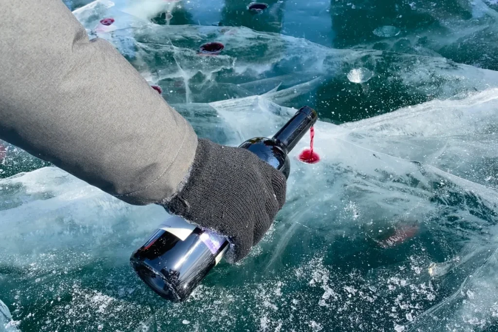 A gloved hand tips a red wine bottle, spilling drops that freeze on a cracked ice surface