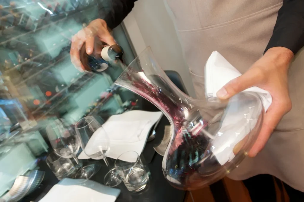 A server pours red wine from a bottle into a wide glass decanter beside empty wine glasses