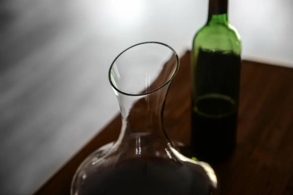 Uncovered glass carafe holding red wine next to an open green wine bottle on a wooden table