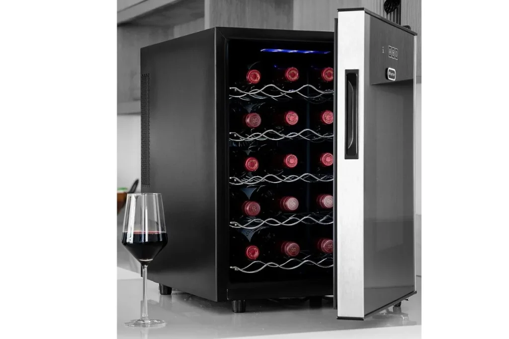 Countertop wine refrigerator with its glass door open, holding rows of red wine bottles beside a filled glass