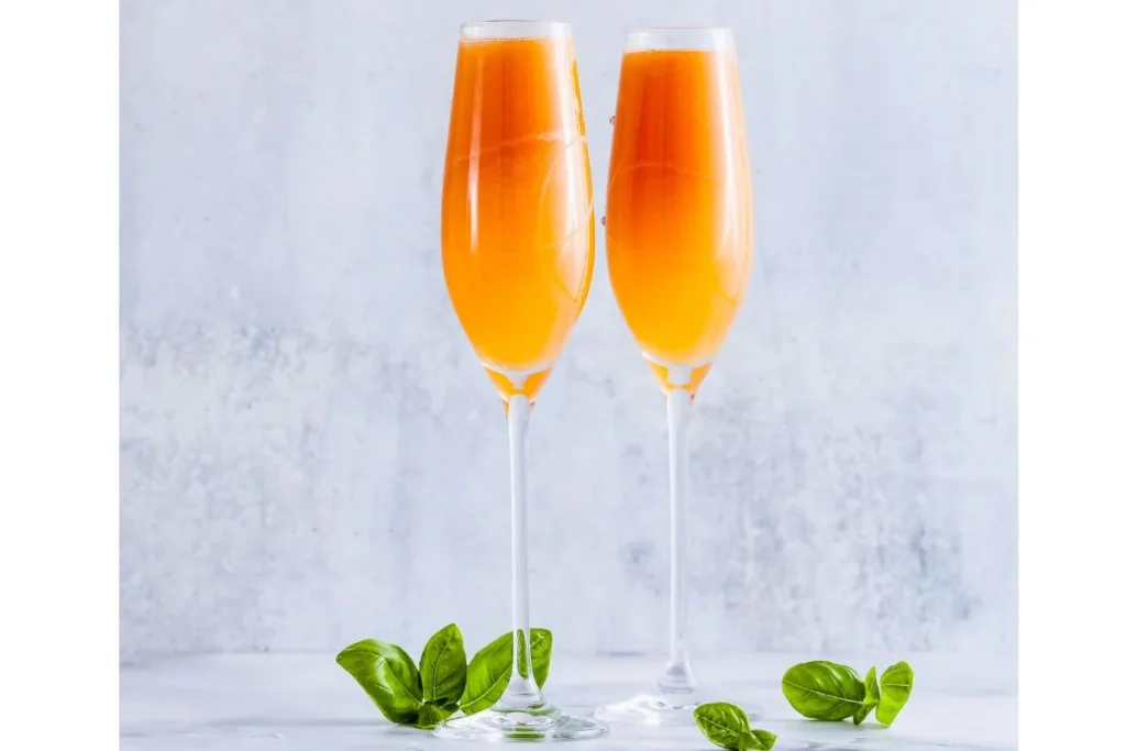 Two champagne flutes filled with an orange sparkling cocktail beside fresh basil leaves