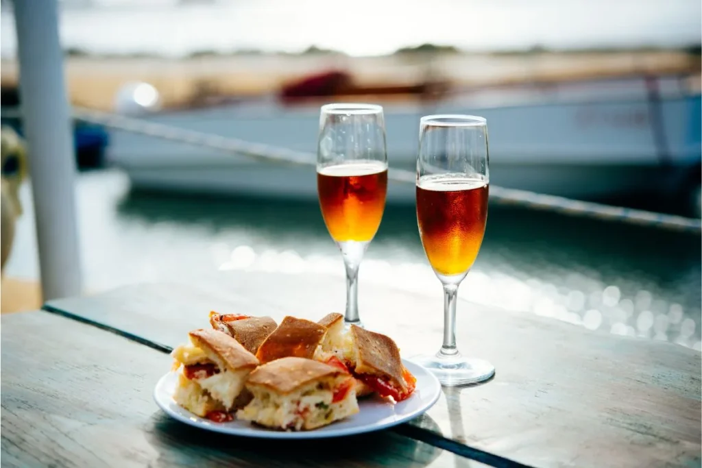 Two flutes of amber fortified wine beside a plate of bread on a waterfront table