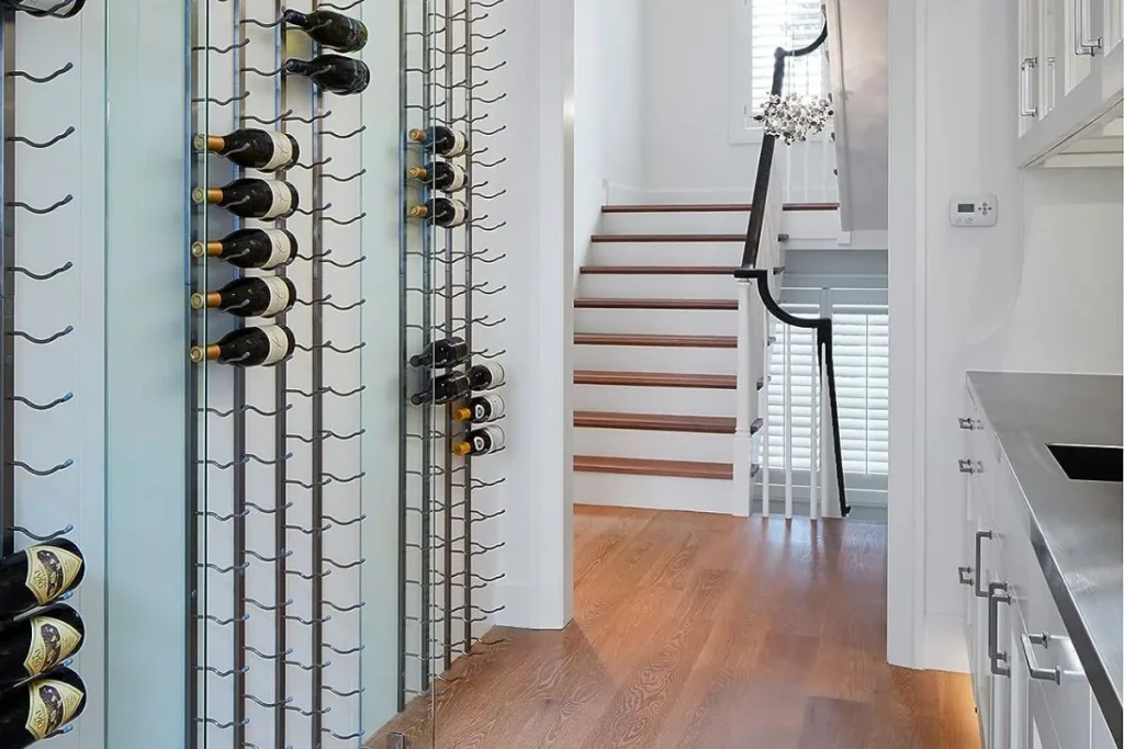 Wall mounted metal wine racks holding bottles horizontally in a narrow hallway near a staircase