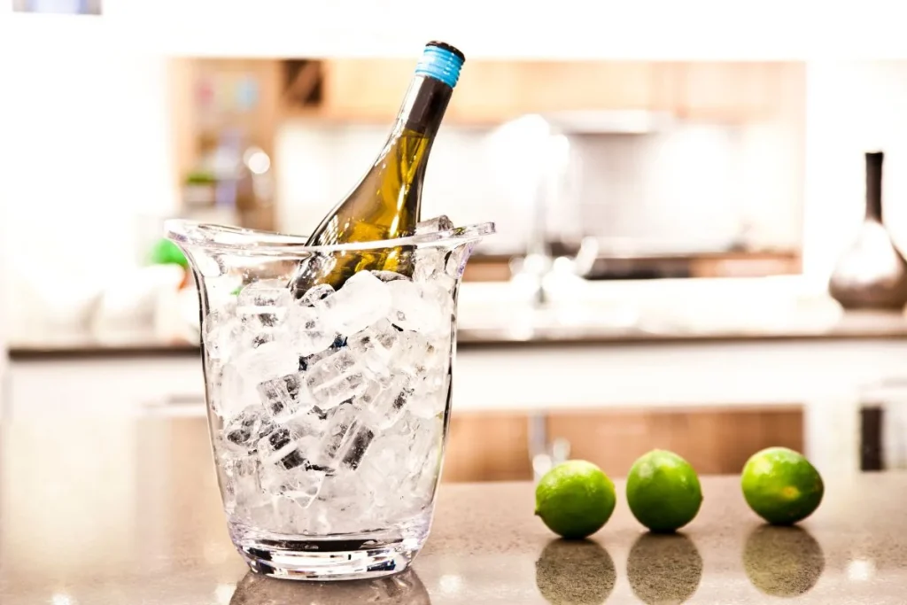 White wine bottle chilling in a glass ice bucket on a kitchen counter beside three limes