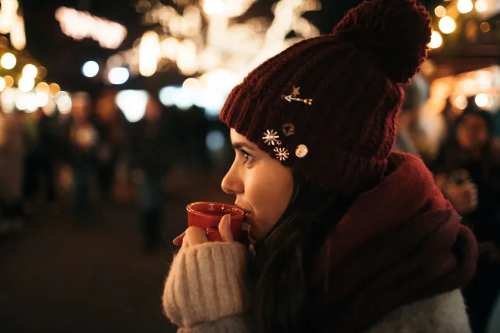 A woman in a knit hat sips mulled wine from a small red mug at a winter market